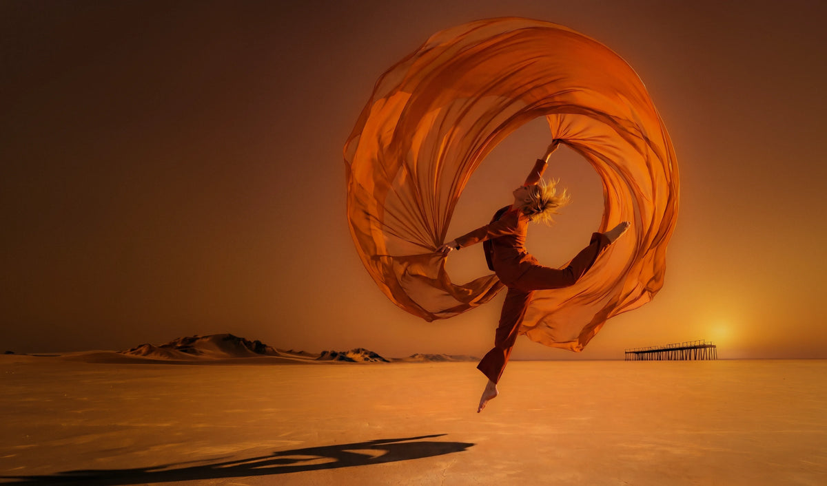A figure turning through desert light inside a sweep of orange silk
