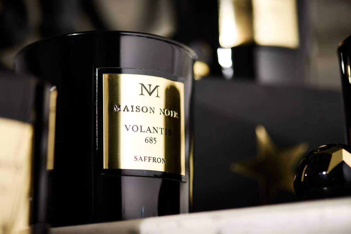 A black glass candle or fragrance vessel with a gold label reading "Maison Noir Volantis 685 Saffron" sits on a white surface with blurred b