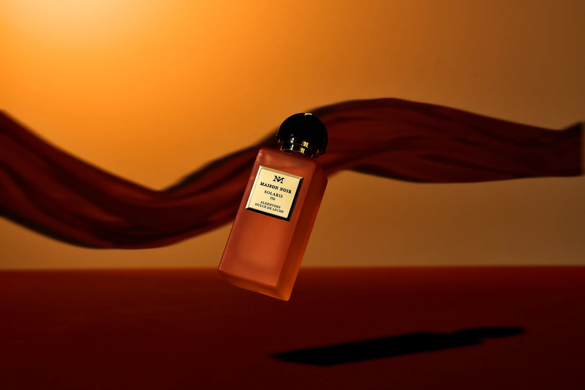 An amber-colored perfume bottle with a black cap and white label sits on a warm-toned surface with draped burgundy fabric in the background