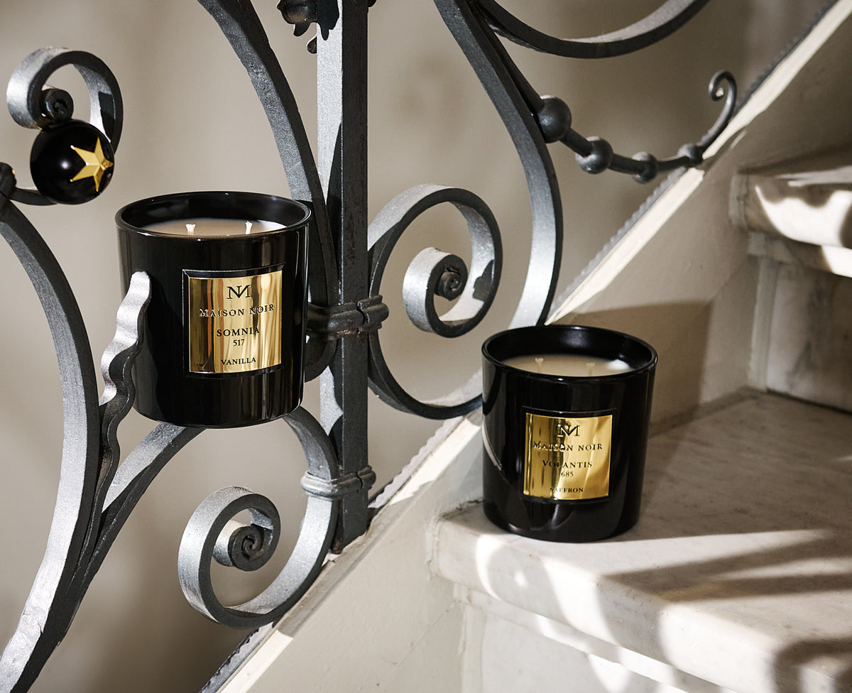 Two black candles with gold labels from Maison Noir sit on an ornate black and silver metal candelabra against a neutral wall