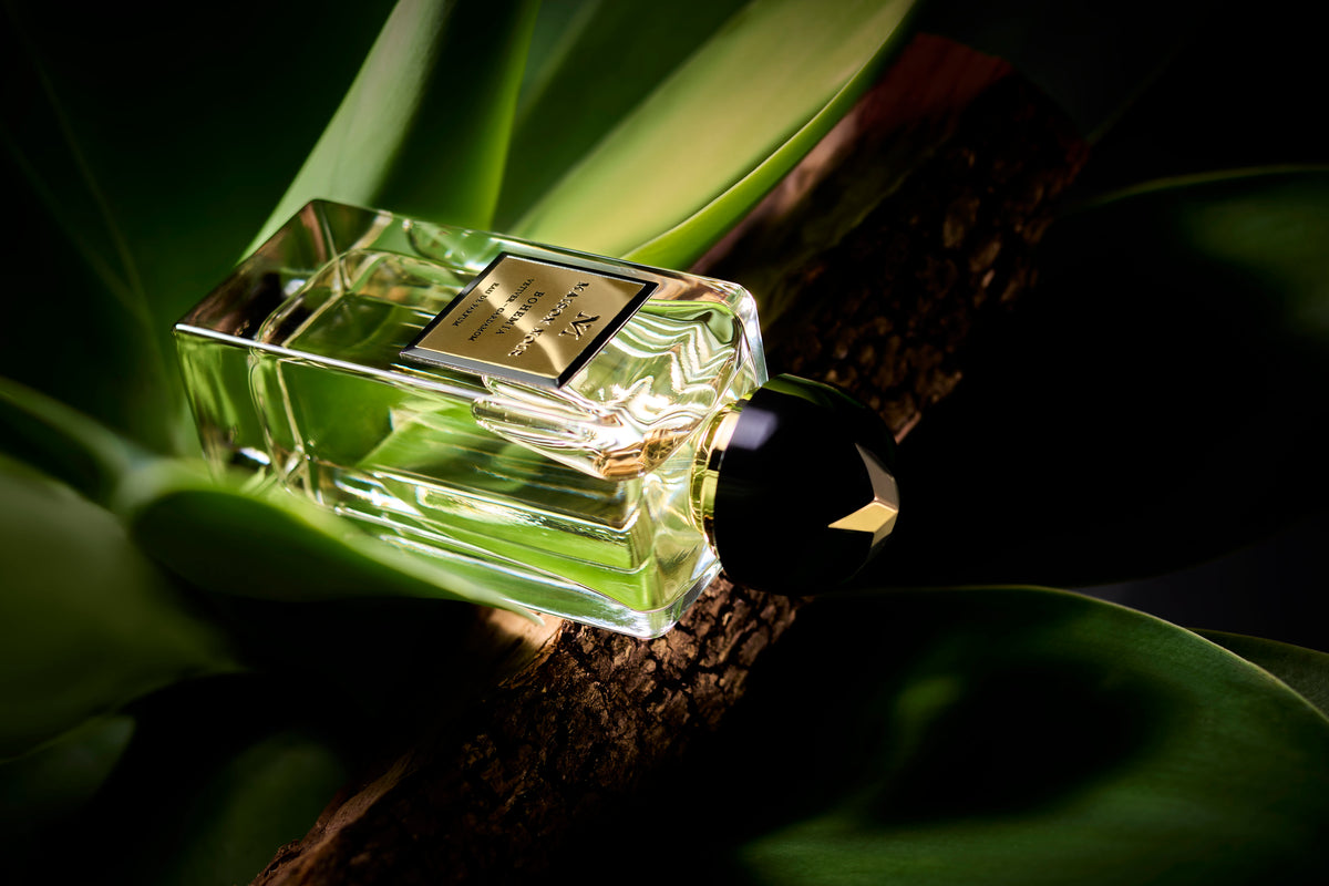 A rectangular glass perfume bottle with pale yellow-green liquid and a metallic label sits on a dark branch surrounded by green foliage