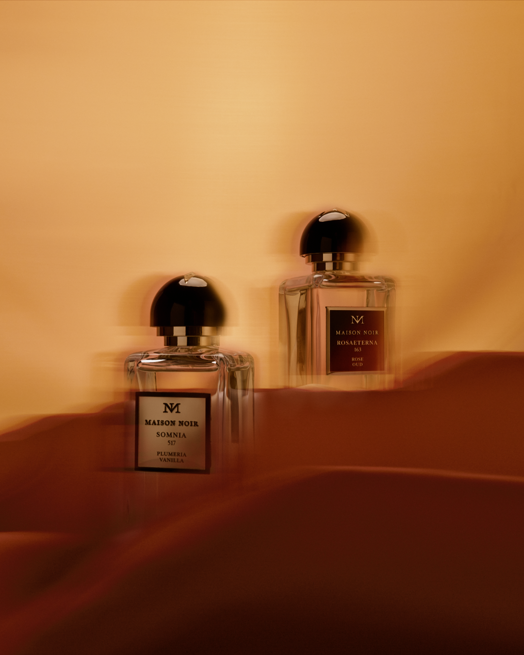 Two Maison Noir perfume bottles with black caps displayed on a warm terracotta surface against a golden background