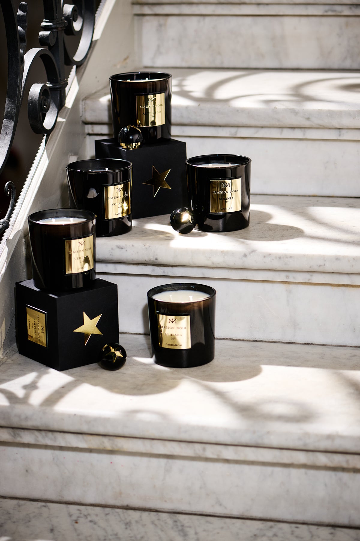 Black glass candles with gold labels arranged on white marble steps in sunlight