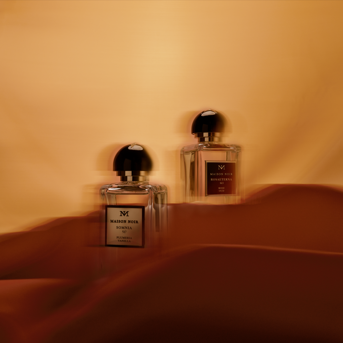 Two Maison Noir perfume bottles with black caps displayed on a warm terracotta-toned surface against a peachy background, casting long shado